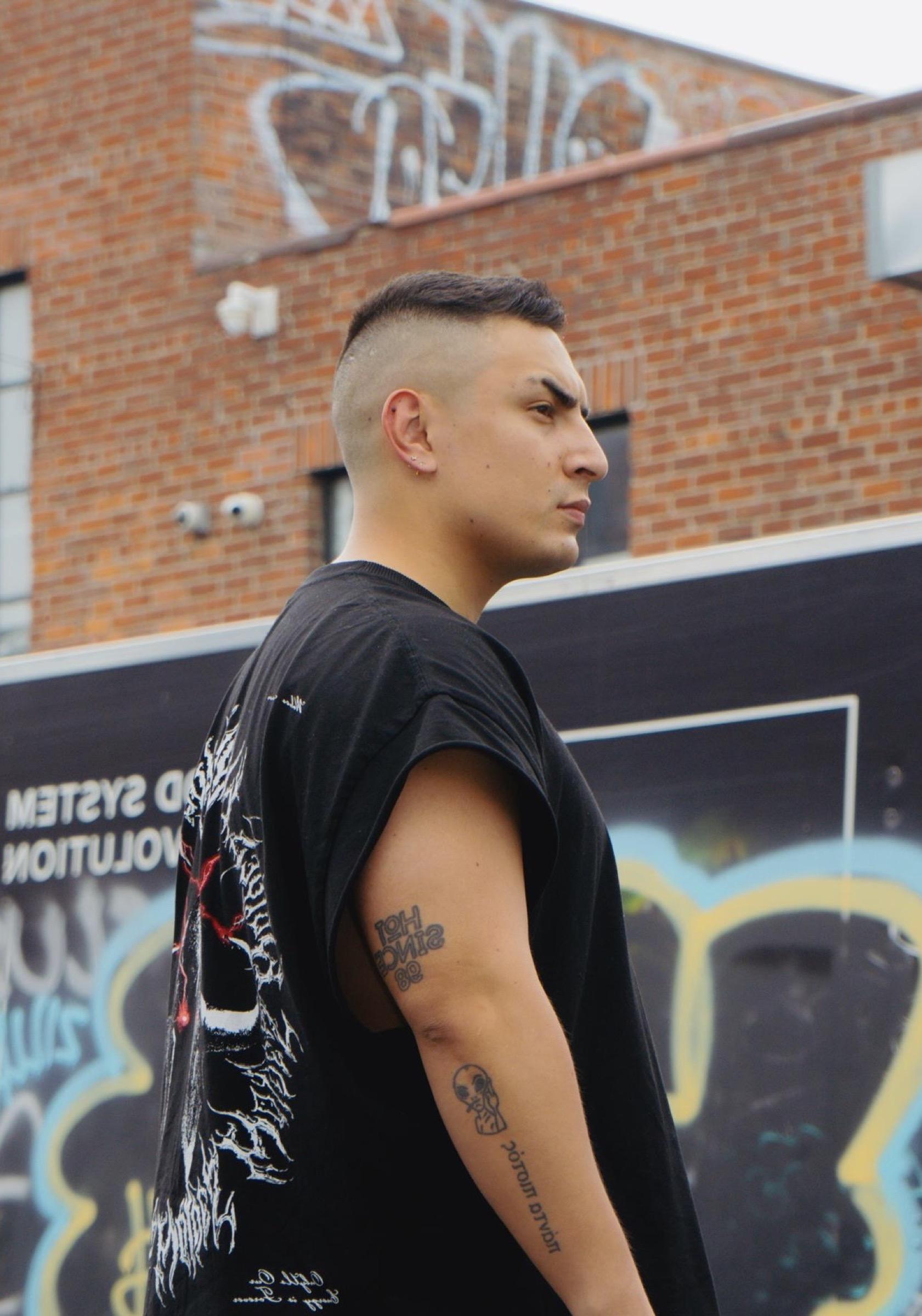 Portrait of Bounc3r in profile in front of a graffiti-covered brick building